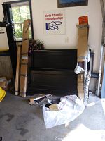 Overview of disassembled queen size wood bed frame components including headboard, footboard, side rails, and slats leaning in a garage with packaging materials and other items around