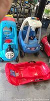 Front view of three children's ride-on toys: a Thomas the Tank Engine car, a Little Tykes police car, and a red sled.