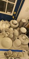 Wide view of stacks of white china dishes, bowls, cups, and serving pieces with silver-gray trim on a covered table near a door.