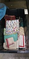 Plastic tub filled with assorted folded linen napkins and tablecloths showing various colors and patterns including floral, checked, and plain fabrics.