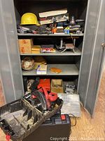 Open metal storage cabinet with multiple tool cases, hardhats, nails, miter box, and small tool accessories, showing organized shelving and some wear on items.