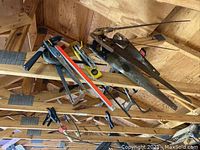 Multiple handsaws, clamps, a level, and measuring yardsticks hung on wooden beams of a garage ceiling.