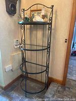 Tall black metal corner shelf with four semi-circular glass shelves and decorative scrollwork frame design, standing near wall and stairs.
