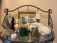 Photo showing a collection of figurines and decorative items on a glass shelf including frog, owl, and ash-made ceramic figures, a white ceramic car coin bank, a small lantern candle holder, framed wall art, and a shell.