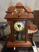 Front view of the wooden mantle clock showing ornate carved top, clock face with Roman numerals, and glass front with visible pendulum.