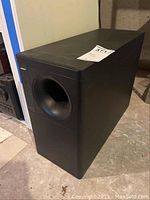 Black Bose Acoustimass 5 Series III subwoofer with front-facing bass port and Bose logo on left front panel, placed on concrete floor with a white sticker on top.