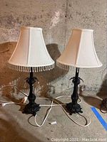 Two matching black table lamps with white fabric shades and dangling bead trim sitting on a concrete basement floor.