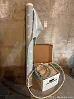 Roll of fabric standing vertically with light blue and white floral pattern, next to a box containing fabric pieces and fabric rings on the floor.