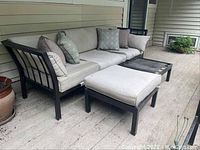 Outdoor patio sofa with metal frame and beige cushions, showing several decorative throw pillows. Sofa placed against exterior house wall on a wood deck. Two ottomans in front, one with cushion missing. Rust visible on metal frames.
