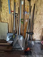 Photo showing variety of yard tools including metal snow shovels, multiple garden rakes with wooden handles, shovels, orange-topped wooden driveway markers/stakes, and the Tanka TBC-220 weed trimmer.