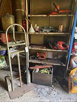 Wide angle showing metal hand truck and shelving unit containing hedge trimmers, chainsaw, and various garden tools.