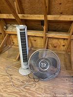 Holmes tower fan model 84001 (FZ25-50D) standing upright next to a metal floor fan in an attic setting. Power cords visible.