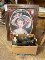 24x30 framed Olympia Beer poster leaning against a wooden structure, with a cardboard box of various glass bottles in front.