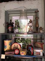 Photo showing a metal shelving unit with 4 lamps on the top shelf and 9 tins of various sizes and designs arranged on shelves beneath it. The background is a white-washed rustic wall with a small window.