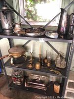 Shelf with metal coffee pot, two pitchers, one round serving tray, smaller pitcher, and various items on lower shelves including candlesticks, cruet set, oil lamp, candlesticks, can opener, hand rake.