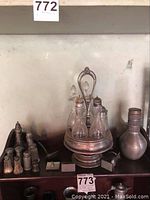 Photo showing a decorative cruet set with glass oil and vinegar bottles on a central metal stand, along with multiple salt and pepper shakers, a tall metal bottle, card holders and napkin ring arranged on a wooden shelf.