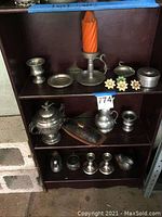 Metal items arranged on brown shelves including candleholders, metal jars, cups, figurines shaped as birds and a duck, metal plates and tins, and flower push pins.