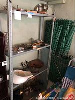 Metal shelving unit with assorted kitchen and household items including a white bed pan on the bottom shelf, metal pot, and various utensils scattered on shelves and in trays.