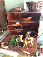 Overview of wooden spice rack shelf showing wooden bowls inside, woven basket on top, and carved camel figure in front, along with drafting tool kit and pastry cutter visible on drop-down surface.