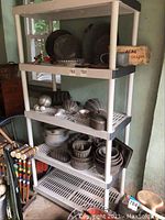 Plastic shelving unit with metal baking pans and molds arranged on three shelves, some spherical metal kitchen accessories, and boxed Acme safety grater on top shelf.