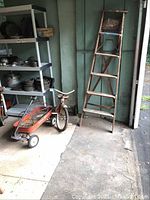 Wide view of garage corner with Radio Flyer tricycle, red Radio Super wagon with wire baskets inside, and wooden step ladder leaning against the wall.