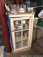 Front view of early American vintage wooden cabinet with glass-paneled door showing wear and scratches. Cabinet contains various dishes seen through glass.