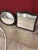 Two early 1900s mirrors leaning against a red wall on concrete floor: one rectangular with dark ornate wood frame, one round with ribbed wood frame.