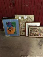 Photo showing all four framed artworks leaning against a wall with visible floor and red background