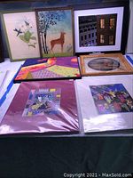 Seven pieces of mixed medium wall art and prints arranged on a table, showing variety of colors, framing and art styles including abstracts and nature themes