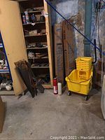 Photo showing corner of basement with wooden shelving containing assorted tools and hardware; yellow industrial mop bucket with wringer and wheels; two cardboard boxes likely containing fluorescent tubes; two fire extinguishers visible on floor; worn concrete walls and floor.