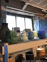 Wide view of two shelves with various pottery including pitchers, vases, and planters in different shapes and colors, all in worn condition.