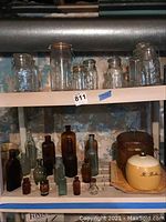 Two shelves showing an assortment of vintage glass jars and bottles with a metal bucket and ceramic container to the right.