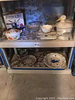 Two shelves of various serving bowls, platters, and dishes including a fondue set box, ceramic duck covered dish, glass bowls, and decorative plates