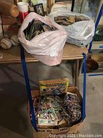 Two plastic bags stuffed with loose baseball cards, sitting on a table, with a partially visible cardboard box beneath containing more cards and sticker album.