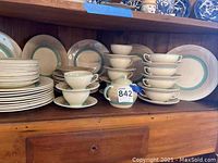 Multiple stacked plates, teacups and saucers arranged on wooden shelf, showing pattern and condition.