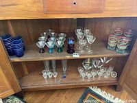 Wide shelf view showing multiple glassware types including colorful tumblers, cordial glasses with colored stems, martini glasses, and decorative glasses with red and gold stripes.