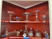 Wide angle photo showing glass cake plates and various glass serving dishes, pitchers, molds, and rolling pin in a red corner shelf.