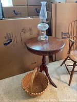 Full view of the round wooden table with hurricane oil lamp on top and basket underneath on carpeted floor with background of moving boxes.