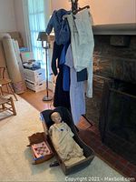 Photo showing multiple handmade clothing pieces hanging on a rack, a vintage doll seated in a small wooden doll cradle, and a larger wooden cradle beside it near a fireplace.