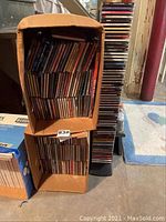 Two large cardboard boxes filled with music CDs in jewel cases, plus a tall CD rack filled with more CDs, stacked vertically.