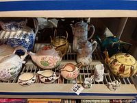 Shelf display of assorted teapots and water pitchers including blue and white ceramics, floral hand-painted pieces, and gold-accented teapot.