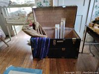 Black vintage trunk opened showing interior with raccoon fur coat, linens, and boxes inside, placed on wood floor near a window.