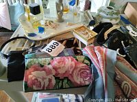 Image shows vintage glass perfume bottles of various sizes and shapes arranged on a floral midcentury ceramic tray alongside floral tapestry and black handbags.