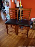 Set of two wooden pub height chairs side-by-side on hardwood floor with orange wall in background. Ladder style backrest and dark cushioned seats.