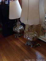 Pair of two matching vintage table lamps, showing bulbous floral etched glass bodies, ornate metal bases, and light fabric lampshades, placed on hardwood floor.