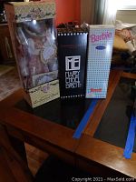 Photo showing three boxed dolls on a table including the Crown porcelain doll with floral box, Mary Engelbreit Toni doll in black box, and Barbie Series 2 Little Debbie doll in white box.