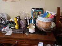 Lot contents on wooden surface including stack of plates, basket, vase, snow globe, candles, flashlights, and packaged wine accessory set.