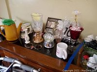 Wide view of yellow ceramic teapot, candle holders, snow globe, mugs, bracelets, pen, small vases, and picture frame on black piano top