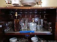Shelf containing assorted small champagne glasses, aperitif glasses, two decanters, and bowls in a wooden cabinet.