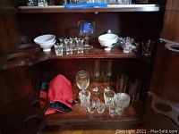 Wide view of wooden cabinet shelf holding assorted glassware, white bowls, a red bottle holder, and insulated carrier.